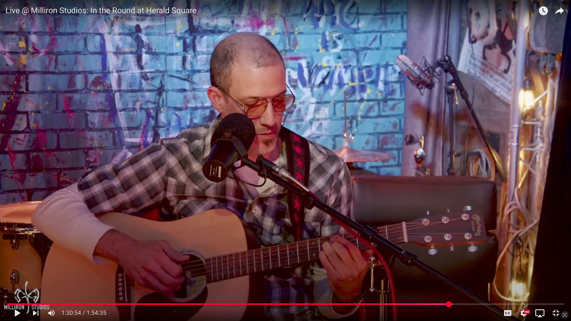 Rob seated, playing guitar, wearing a black and white plaid shirt, in front of a blue graffiti tagged brick wall