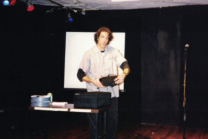 Rob with shaggy hair, on stage in a tiny theater holding a slide projector looking at the camera. Next to him is a table with 2 slide carousels and a black case